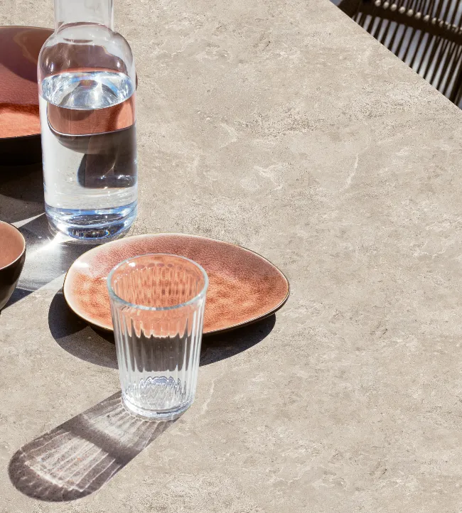 Close-up of the Kodo Dining Table by Vincent Sheppard, featuring a Sabbia Panna ceramic stone top, styled with glassware and copper-toned plates, with woven dining chairs in the background.